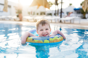 Fototapeta na wymiar Little three years old kid boy swimming and splashing in swimming pool in swimming circle. Smiling and laughing Toddler having fun at summer vacation, leisure activity