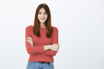 Indoor shot of positive charming european woman with brown hair, holding hands crossed over chest and smiling broadly, feeling uncomfortable but polite over gray background, talking with client