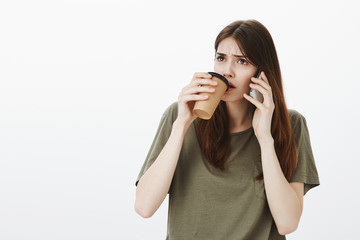 Girl is intense and uncomfortable, carefully listening terrifying story of friend during mobile talk. Portrait of focused worried good-looking woman, sipping coffee and holding smartphone near ear