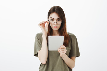 Attractive female woman with confident expression, knows everything. Focused good-looking brunette with trendy glasses, holding hand on rim and staring suspiciously at camera, holding digital tablet