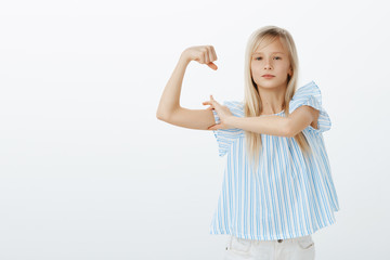 Maybe I am small but strong. Portrait of proud confident young blond girl in stylish blouse, raising hand and showing muscles, talking about strength, feeling self-assured and pleased over gray wall