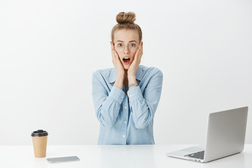 Shocked impressed female coworker in glasses, holding palms on face while hearing amazing rumor or gossip, talking with colleague, sitting in office near laptop and smartphone, drinking coffee