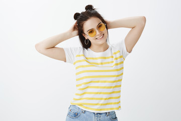Time to have some fun. Carefree confident and relaxed stylish feminine woman in sunglasses and striped t-shirt, holding hands behind head and smilng joyfully while posing over gray background