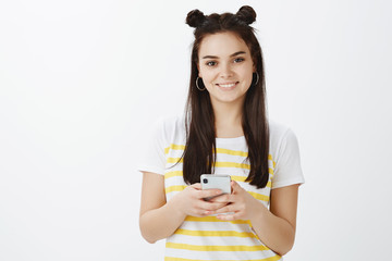 Waist-up shot of modern good-looking happy woman with cute hairbuns, holding smartphone and smiling joyfully while gazing at camera over gray background, using new gadget for first time