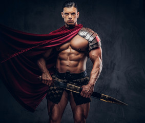 Brutal ancient Greek warrior with a muscular body in battle equipment posing on dark background.