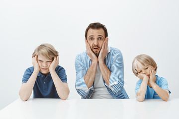 How they quickly grew up. Portrait of shocked anxious european father sitting with sons, holding hands on face and dropping jaw, feeling nervous while siblings are bored and careless over gray wall