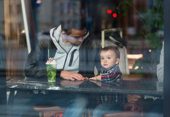 Father and son sitting at cafe. Portrait shot through glass from outside. Father in leather jacket, son in checkered shirt. Hipster urban family leisure.