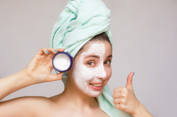 Woman with face cream on her face demonstrating a jar with white facial cream and showing a thumbs up.