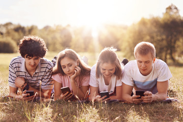 Four friends focused in their smart phones, enjoy leisure time on nature, lie on green grass, share photos after picnic in their social networks, pose against sunshine. Teenagers, technology