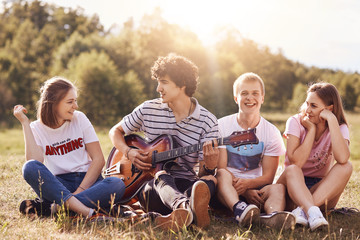 Happy young friends sing songs to guitar, have joy together, recreat outdoor, sit on green grass. Curly handsome male teenager plays guitar, enteratins his companions, enjoys summer warm day.