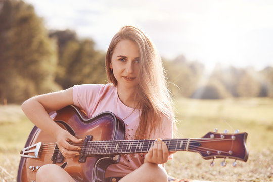 People, talents, music concept. Attractive young female teenager plays music instrument, likes guitar, looks happily directly at camera, dressed in casual t shirt, poses outdoor against sunshine