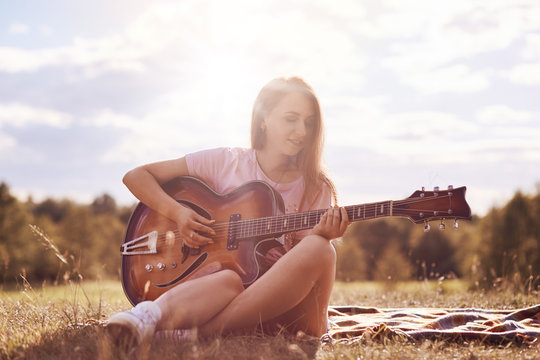 Attractive young female guitarist uses music instrument, plays guitar, sings song, produces wonderful sound or melody, sits crossed legs on plaid, poses against blue sky and sunshine background