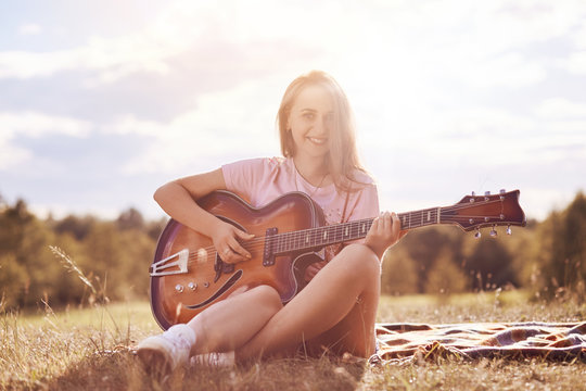 Horizontal shot of good looking European female with happy expression plays guitar, looks confidently at camera, sits crossed legs on plaid, poses against sky with white clouds. People, music concept