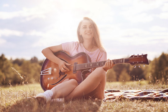 Beautiful talented young female musician plays acoustic guitar, has gentle smile on face, sits on green grass of meadow, has joyful expression, sings her favourite song, enjoys sunny day outside