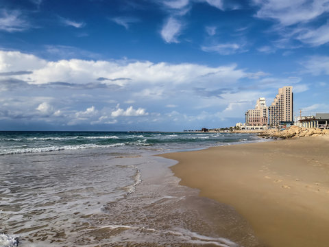 A view of a lonely romantic beach with a rolling wave on a sand shore and  high buildings on the horizon under the blue sky with white clouds. The photo was shot in a public beach of Haifa, Israel. 