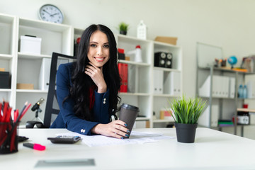 A beautiful young girl is holding a glass with coffee, sitting on a chair in the office at the table.