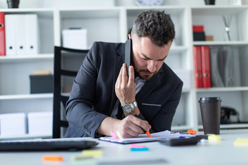 A man is sitting at the desk at the office, talking on the phone and writing a marker in the document.