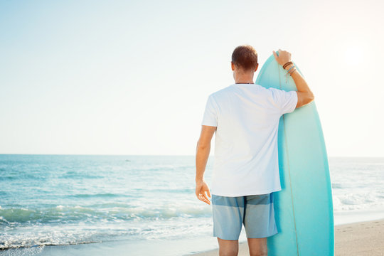 Portrait of happy surfer in hawaiian t-shirt walking with surf board on the beach at sunset