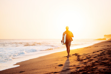 Obraz premium Portrait of happy surfer in hawaiian t-shirt walking with surf board on the beach at sunset