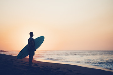 Portrait of happy surfer in hawaiian t-shirt walking with surf board on the beach at sunset