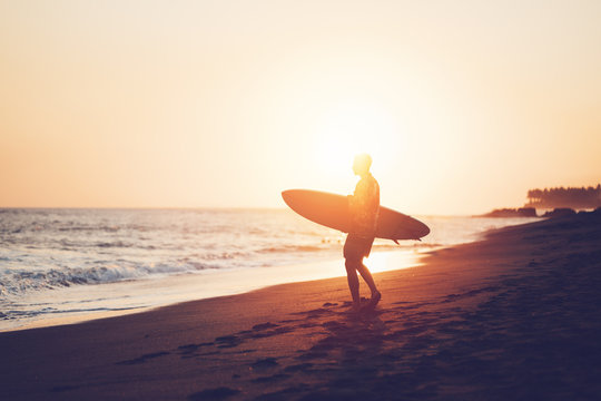 Portrait of happy surfer in hawaiian t-shirt walking with surf board on the beach at sunset