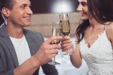 I have wonderful toast. Cheerful young man and woman are clinking champagne glasses and smiling. They are looking at each other with love. Bride and groom are sitting on bed 