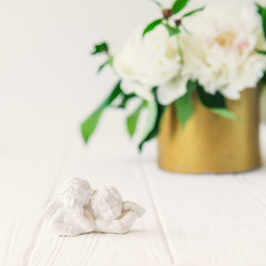 Close up statuette of two antique little lovely angels of the gypsum on the white wooden table with bouquet of white peonies in vintage bowl. Love background. Square card. Copy space, selective focus.