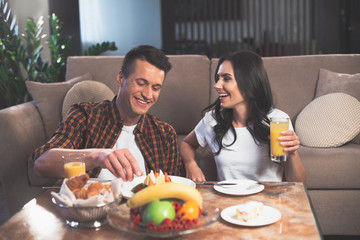 Excited young lovers are eating sweet food with enjoyment. They are talking and laughing while relaxing in living room