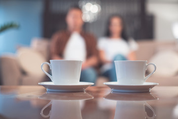 Low angle focus on white cups of tea on small plates on desk. Married couple is relaxing on sofa on background