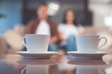 Focus on white mugs of hot beverage on table. Cheerful young man and woman are sitting on couch on background. Home coziness concept 