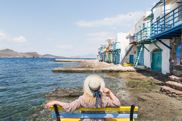 Young blonde girl sitting on a bench overlooking colourfull old houses in fishermen town of Klima on Milos island, Greece