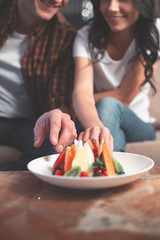 Low angle close up of male and female arms taking fresh fruits from plate. Happy couple is smiling while sitting on couch near the table. Focus on dessert 