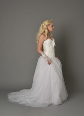 full length portrait of blonde  girl wearing white fantasy gown. standing pose in side profile, grey studio background.