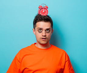 Young handsome man in orange t-shirt with red alarm clock. Studio image on blue background