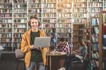 Portrait of beaming male working with digital device while locating in athenaeum. Smiling students learning there