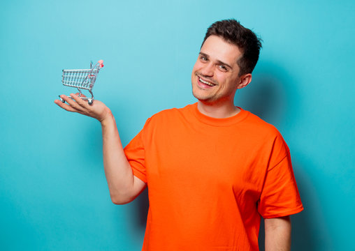 Young handsome man in orange t-shirt with shopping cart. Studio image on blue background