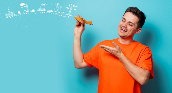 Young handsome man in orange t-shirt with wooden airplane and dreaming travel route. Studio image on blue background