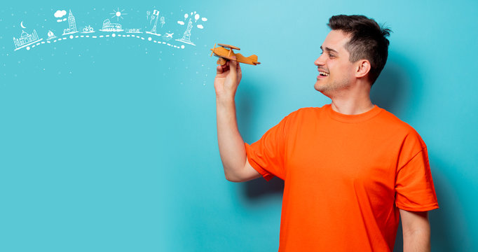 Young Handsome Man In Orange T-shirt With Wooden Airplane And Dreaming Travel Route. Studio Image On Blue Background