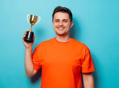 Young handsome man in orange t-shirt with golden cup. Studio image on blue background