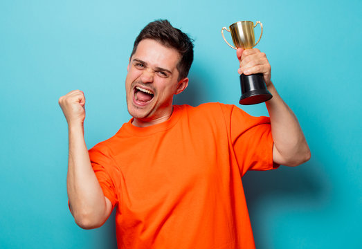 Young handsome man in orange t-shirt with golden cup. Studio image on blue background