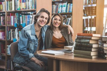 Portrait of cheerful man embracing outgoing female while sitting at table in athenaeum. They using gadget