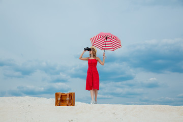 Young woman in red dress with umbrella and suitcase looking in binoculars on the beach. Travel concept image on sand