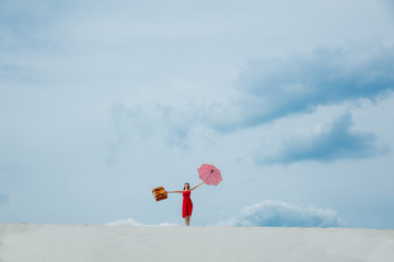 Fototapeta na wymiar Young woman in red dress with umbrella and suitcase on the beach. Travel concept image on sand