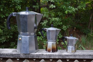 Oversized, 6 cup and 3 cup moka coffee maker outside on a railing made of wood.