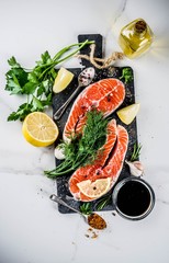 Raw salmon fish steaks with lemon, herbs, olive oil, ready for grill, slate cutting board, white marble background copy space above