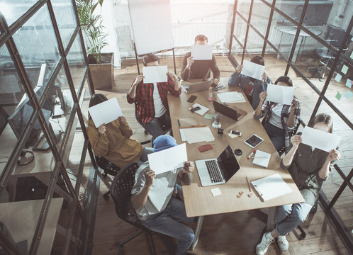 Read this. Full length top view of young trendy coworkers are sitting around desk with different modern devices. They are laboring in office and covering their faces while holding sheets of paper