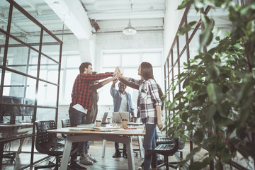 We are best team. Low angle full length of successful young friendly colleagues are standing around table in office and holding hands together. They are showing their togetherness and partnership