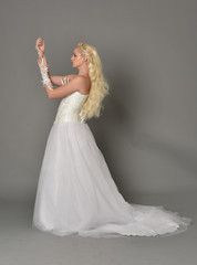 full length portrait of blonde  girl wearing white fantasy gown. standing pose in side profile, grey studio background.