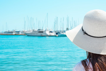 Attractive Young Caucasian Woman with Long Hair in Hat Stands at Beach Looks at Yachts Sailing Boats Moored in Marina. Turquoise Sea Blue Sky Sunny Summer Day. Vacation Wanderlust Romantic