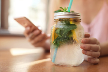 Young woman using mobile phone while drinking tasty natural lemonade at table in cafe. Detox drink - obrazy, fototapety, plakaty